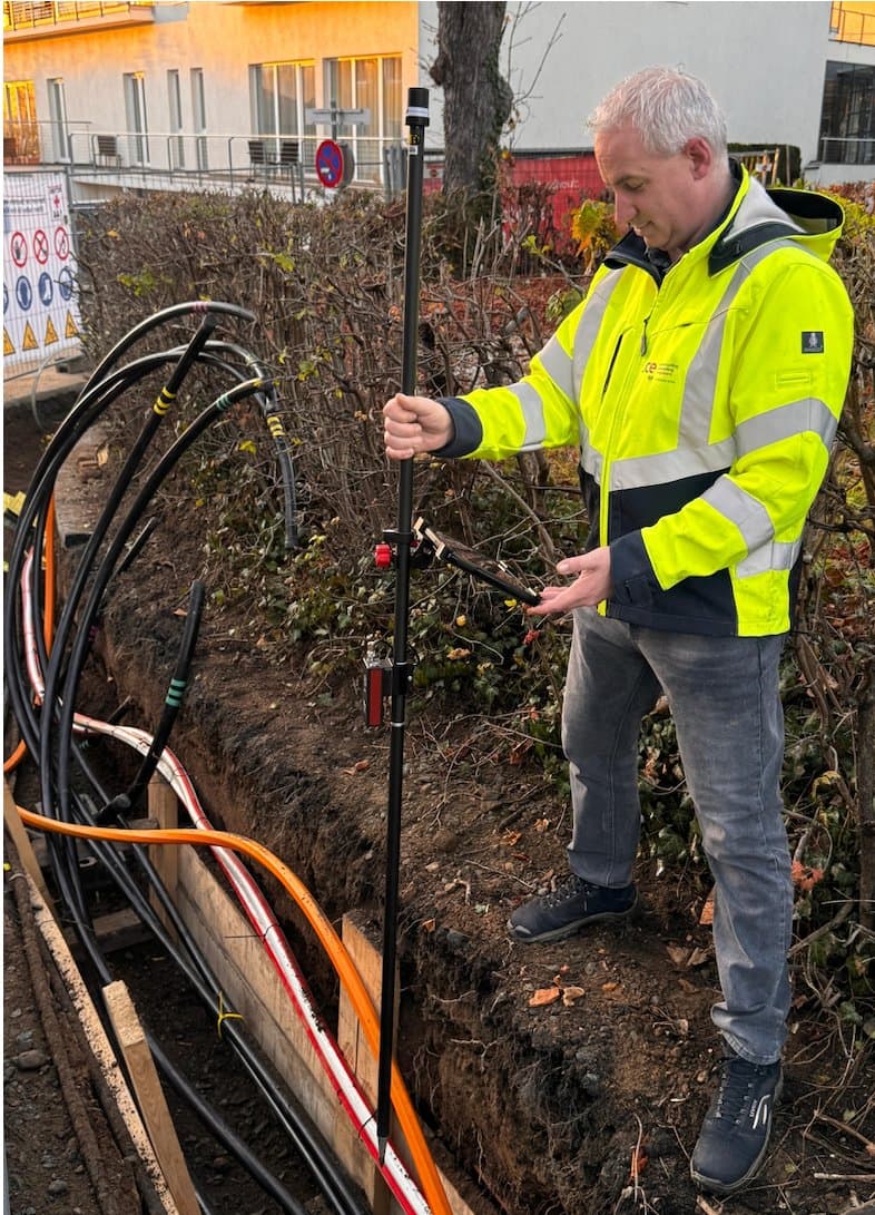 A site engineer measures an open cable trench with an RTK pole and a tablet